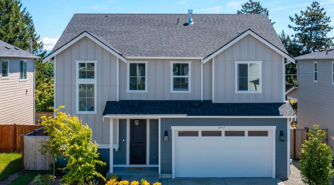 Front view of a modern gray two-story house with a double-gabled roof, two large windows, a white-framed front door, a white garage door, and a landscaped yard under a bright blue sky. Address 2407 is visible on the garage door.