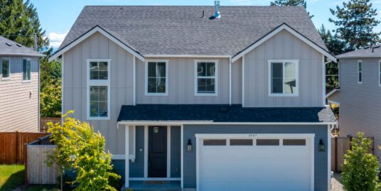 Front view of a modern gray two-story house with a double-gabled roof, two large windows, a white-framed front door, a white garage door, and a landscaped yard under a bright blue sky. Address 2407 is visible on the garage door.